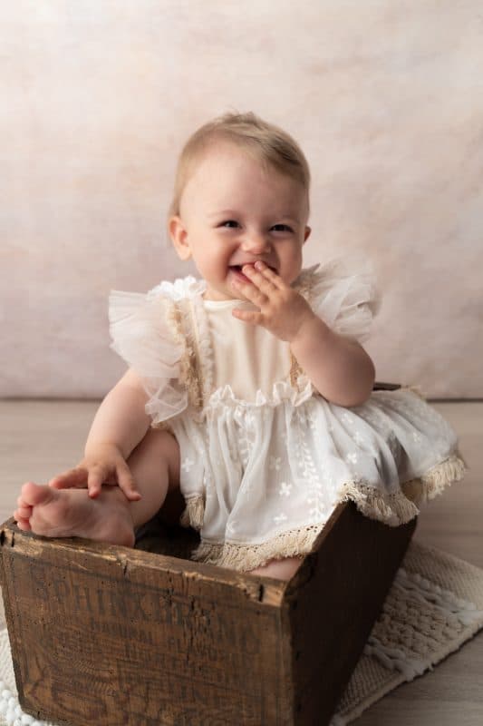 Un bébé souriant, vêtu d'une robe blanche à  froufrous, est assis dans une boîte en bois. Le bébé se couvre la bouche d'une main et a les pieds nus posés sur le bord de la boîte. Le fond est doux et aux tons neutres | Cette photo de bébé est réalisée par Tribe Photography | Gaëlle Massart