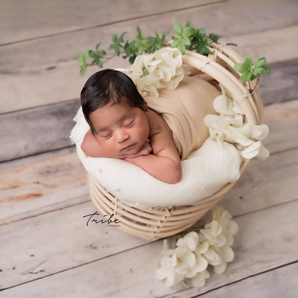 Un nouveau-né, enveloppé dans un tissu beige, dort paisiblement dans un panier en osier orné de fleurs blanches et de feuilles vertes. Le panier est posé sur un parquet rustique en bois et le bébé repose sur une couverture blanche. Cette ravissante photo naissance bébé est marquée du logo Tribe Photography | Cette photo de naissance garà§on est réalisée par Tribe Photography | Gaëlle Massart