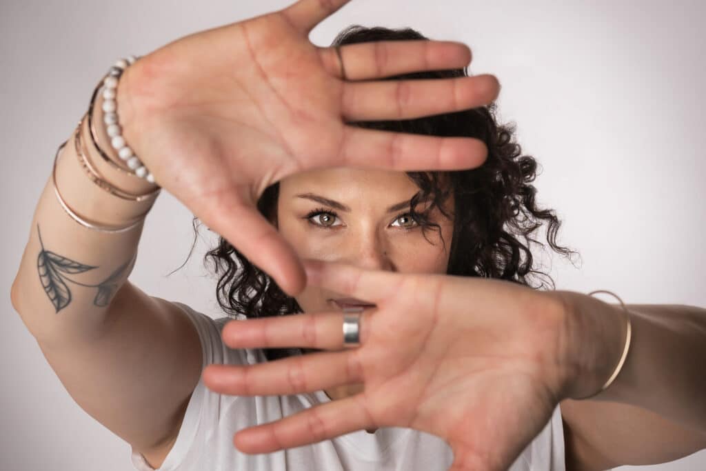 Une femme aux cheveux bouclés et aux multiples bracelets aux poignets tient ses mains devant son visage, encadrant ses yeux comme un mannequin. Elle porte une chemise blanche et ont un tatouage visible sur un bras. La photo, parfaite pour un livre photo, a un fond neutre | Cette séance photo pour book mannequin est réalisée par Tribe Photography | Gaëlle Massart Photographe book mannequin