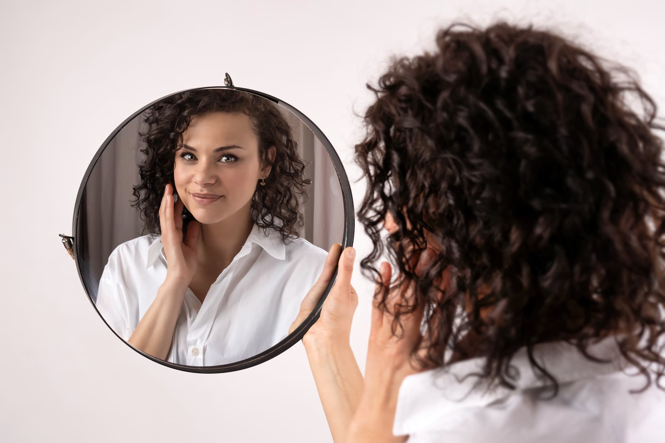 Une femme aux cheveux bouclés regarde son reflet dans un miroir rond, touchant son visage. Elle porte une blouse blanche et sourit légèrement, comme si elle se préparait pour un casting photo. Le fond est uni et de couleur claire | Cette séance photo pour casting est réalisée par Tribe Photography | Gaëlle Massart Photographe