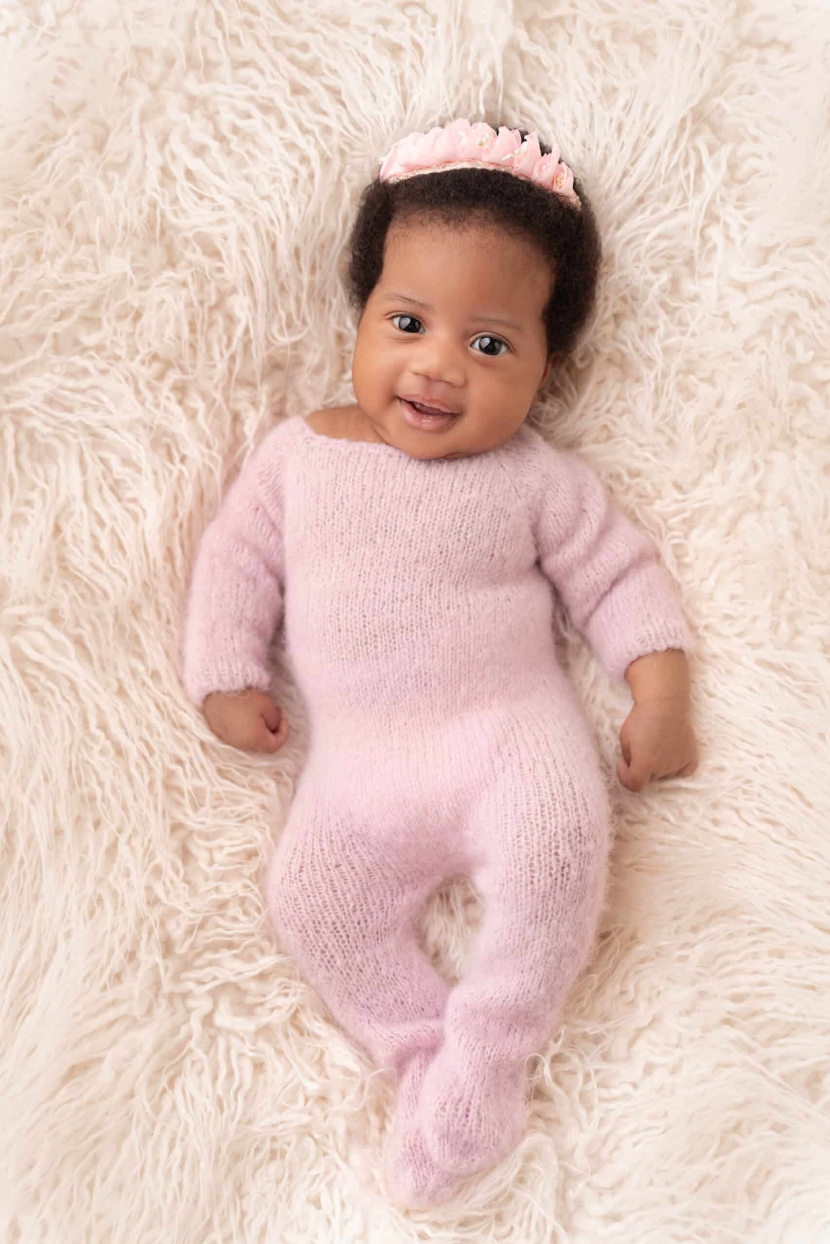 Un bébé, vêtu d'une combinaison tricotée rose tendre et d'un bandeau fleuri, est allongé sur une couverture blanche moelleuse tout en souriant et en regardant l'appareil photo pendant sa séance photo de naissance | Ce shooting photo naissance est réalisé par Tribe Photography | Gaelle Massart Photographe