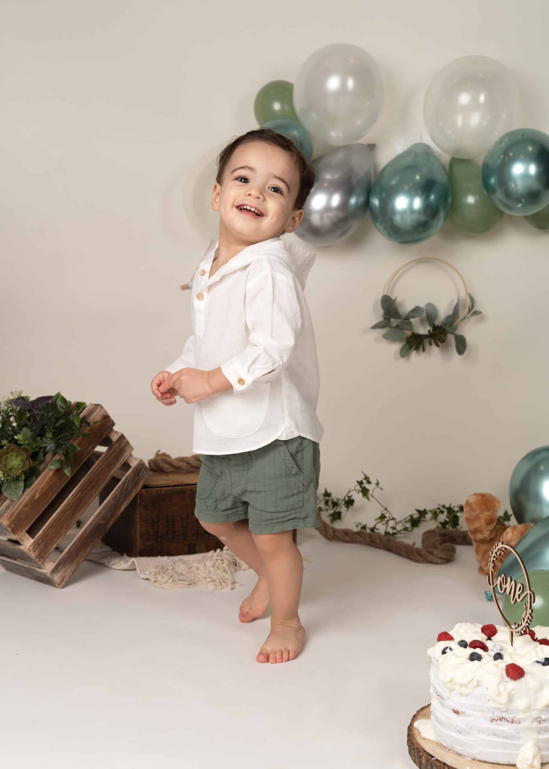 Un enfant en bas âge joyeux avec un grand sourire se tient pieds nus devant un décor simple et festif avec des ballons bleu sarcelle et argent, une caisse en bois et de la verdure. L'enfant porte une chemise blanche et un short vert, parfait pour cette séance photo bébé. Un petit gâteau avec une décoration « One » est visible au premier plan | Cette séance photo bébé est réalisée par Tribe Photography | Gaelle Massart Photographe