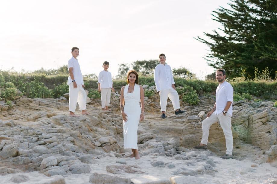 Une famille de cinq personnes, toutes vêtues de blanc, se tient sur un terrain rocheux près de la plage. Quatre hommes et une femme posent parmi les rochers avec de la verdure et des arbres en arrière-plan. Le ciel semble clair avec la lumière naturelle du soleil | Cette photo de famille est réalisée par Tribe Photography | Gaëlle Massart
