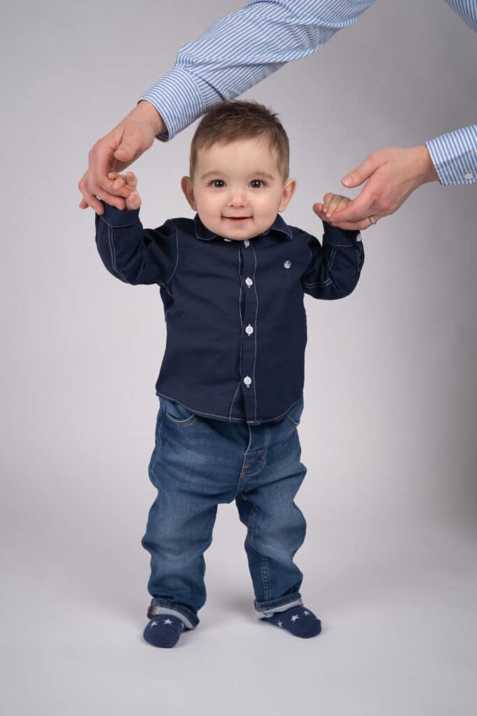 Un bébé souriant se tient debout tout en étant doucement tenu sous les bras par les mains d'un adulte lors d'une séance photo bébé. Le bébé est habillé d'une chemise bleu marine, d'un jean bleu et de chaussettes à motifs. Le fond est uni gris clair | Ce shooting photo bébé est réalisée par Tribe Photography | Gaelle Massart Photographe