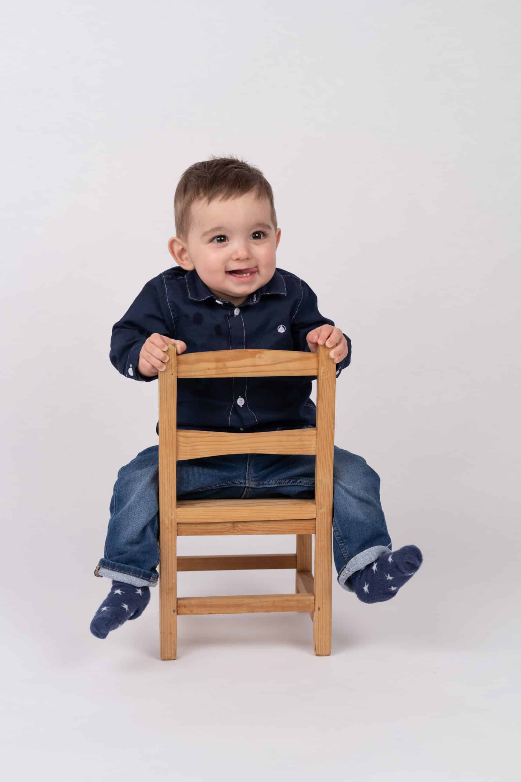 Un bébé aux cheveux bruns courts est assis sur une petite chaise en bois. Le bébé, vêtu d'une chemise bleu foncé à boutons, d'un jean en denim et de chaussettes à motifs étoilés, sourit et regarde légèrement de côté. L'arrière-plan est blanc uni, capturant l'innocence du moment dans cette photo de bébé magnifiquement prise | Ce shooting photo bébé est réalisée par Tribe Photography | Gaelle Massart Photographe