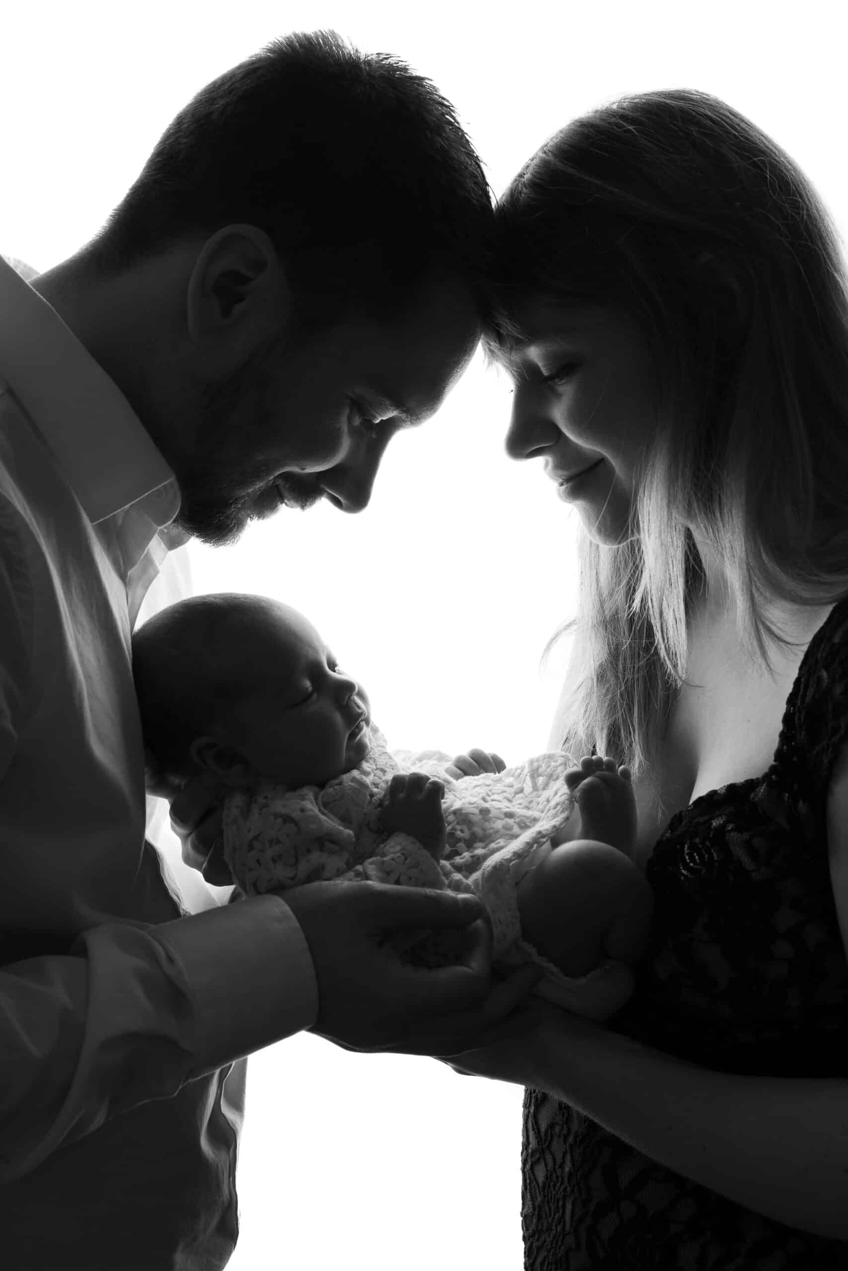 Une photo en noir et blanc montre un couple souriant à leur nouveau-né, qu’ils tiennent doucement entre eux. La tête du père touche celle de la mère dans une tendre démonstration d'affection, et le visage du bébé est paisible et calme : un moment parfait capturé lors de leur séance photo de naissance | Ce shooting photo naissance est réalisé par Tribe Photography | Gaelle Massart Photographe