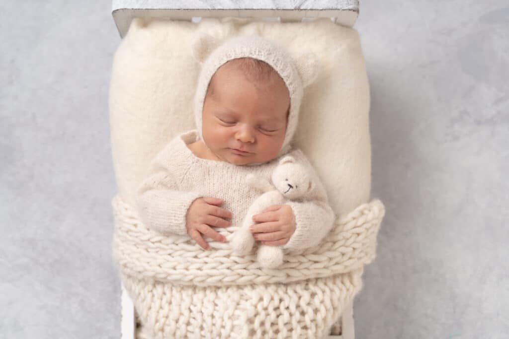 Un nouveau-né dort paisiblement dans un ensemble blanc et confortable, portant un chapeau moelleux en forme d'oreilles d'ours. Le bébé est vêtu de vêtements doux en tricot blanc et tient un petit ours en peluche. Le fond gris clair ajoute à l'atmosphère sereine et réconfortante de cette belle séance photo naissance | Ce shooting photo naissance est réalisé par Tribe Photography | Gaelle Massart Photographe