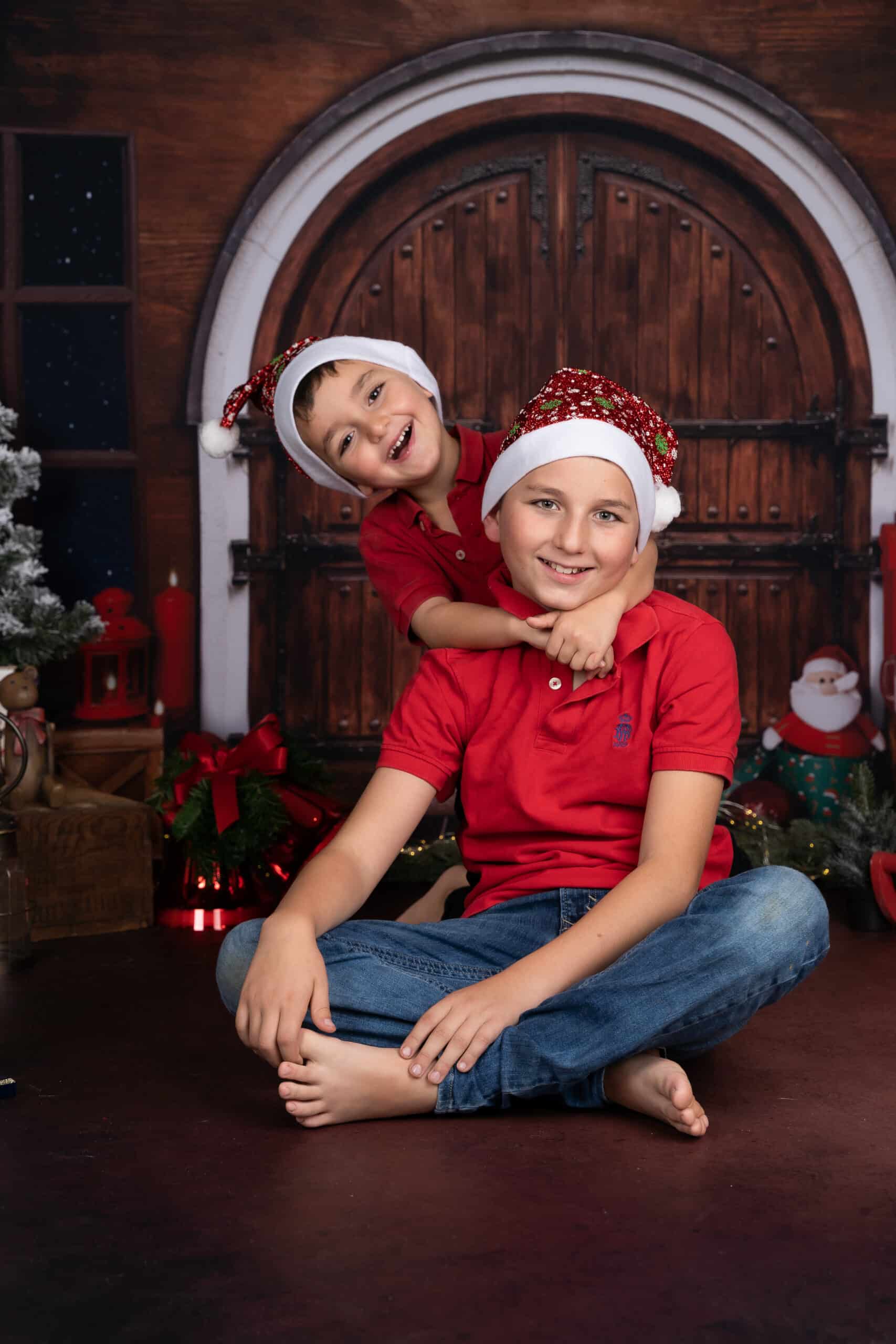 Deux enfants, vêtus de chemises rouges et de chapeaux de Père Noël, posent devant une porte cintrée en bois ornée de décorations festives. Un enfant sourit depuis le sol tandis que l'autre s'enlace de manière ludique par derrière ; tous deux rayonnent de joie. Un sapin de Noël et des cadeaux créent le décor parfait pour cette séance photo Noël | Ce shooting photo Noël famille est réalisée par Tribe Photography | Gaelle Massart Photographe