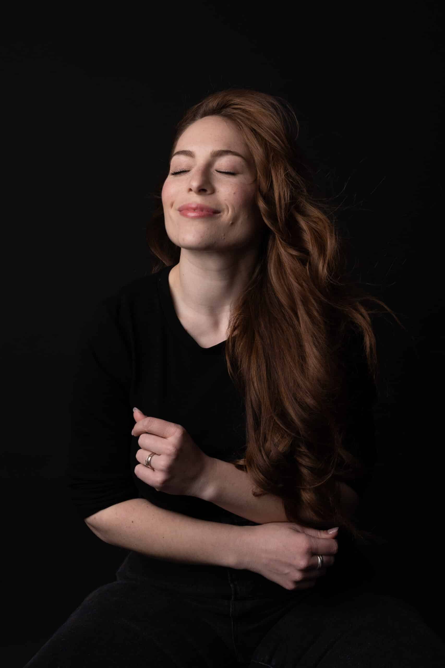 Une femme aux longs cheveux roux flottants est assise sur un fond noir dans ce portrait photo saisissant. Vêtue d'un haut noir, elle sourit doucement, les yeux fermés, paraissant sereine et contente. Une main est légèrement serrée tandis que l'autre repose lâchement sur ses genoux, capturant un moment de casting paisible | Cette séance photo professionnelle est réalisée par Tribe Photography | Gaëlle Massart Photographe