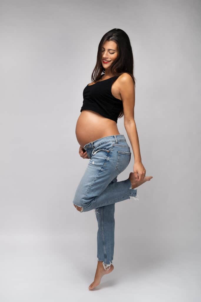 Un photographe grossesse capture une femme enceinte aux longs cheveux noirs, vêtue d'un débardeur noir et d'un jean bleu, alors qu'elle pose sur une jambe sur un fond neutre. Elle sourit doucement avec sa main droite posée sur son ventre et sa jambe gauche pliée derrière elle | Cette photo de grossesse est réalisée par Tribe Photography | Gaëlle Massart