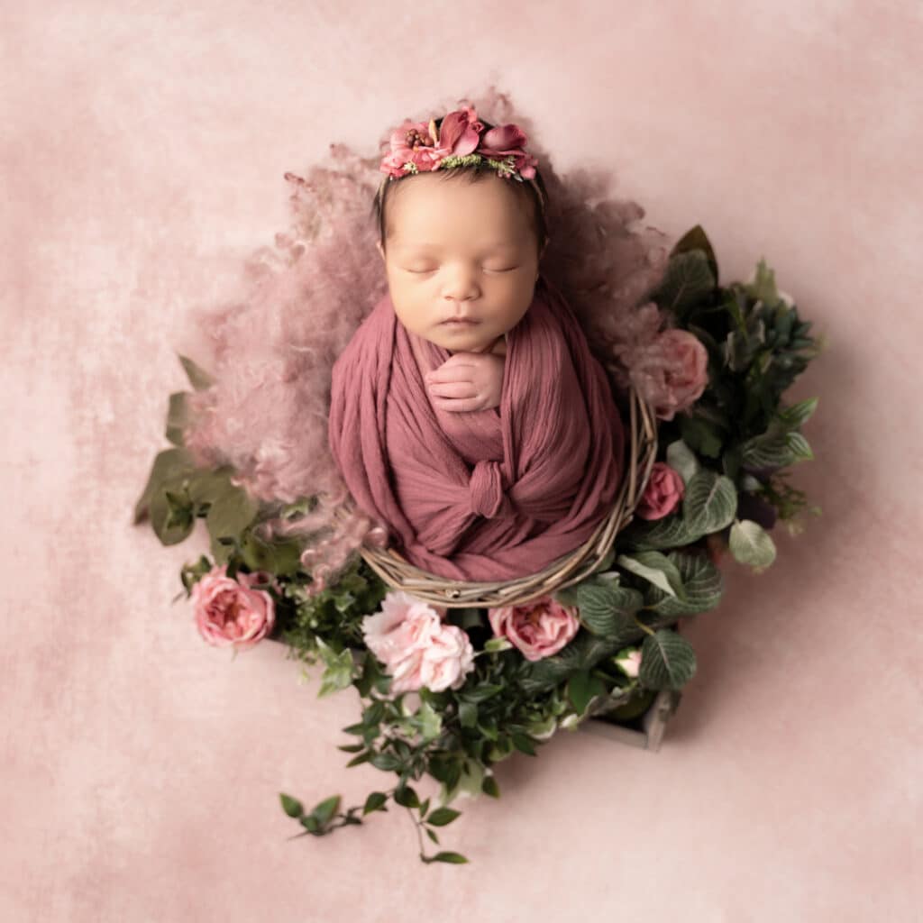 Un nouveau-né dort paisiblement, emmailloté dans une couverture mauve et orné d'un bandeau fleuri assorti. Le bébé est niché dans un panier entouré de textures douces et moelleuses et de roses roses aux feuilles vertes, le tout sur un fond rose clair lors de la parfaite séance photo naissance | Ce shooting photo naissance est réalisé par Tribe Photography | Gaelle Massart Photographe