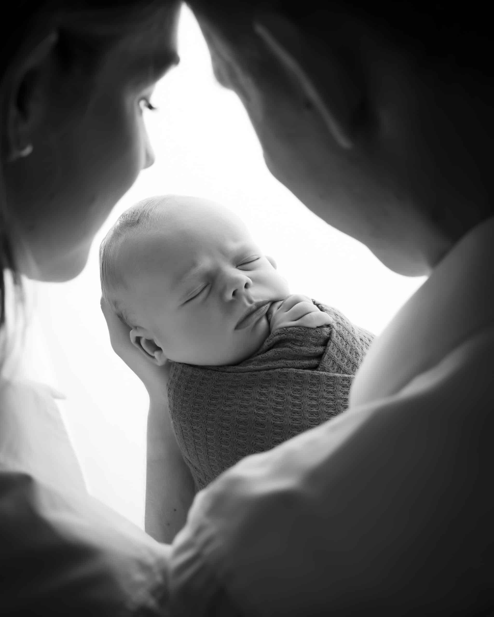 Une photo en noir et blanc d'une séance photo naissance montre un bébé enveloppé dans une couverture, bercé par deux adultes dont les visages sont flous. Le bébé dort paisiblement, la main près de la bouche, et les doux regards des adultes sont dirigés vers le bébé | Ce shooting photo naissance est réalisé par Tribe Photography | Gaelle Massart Photographe