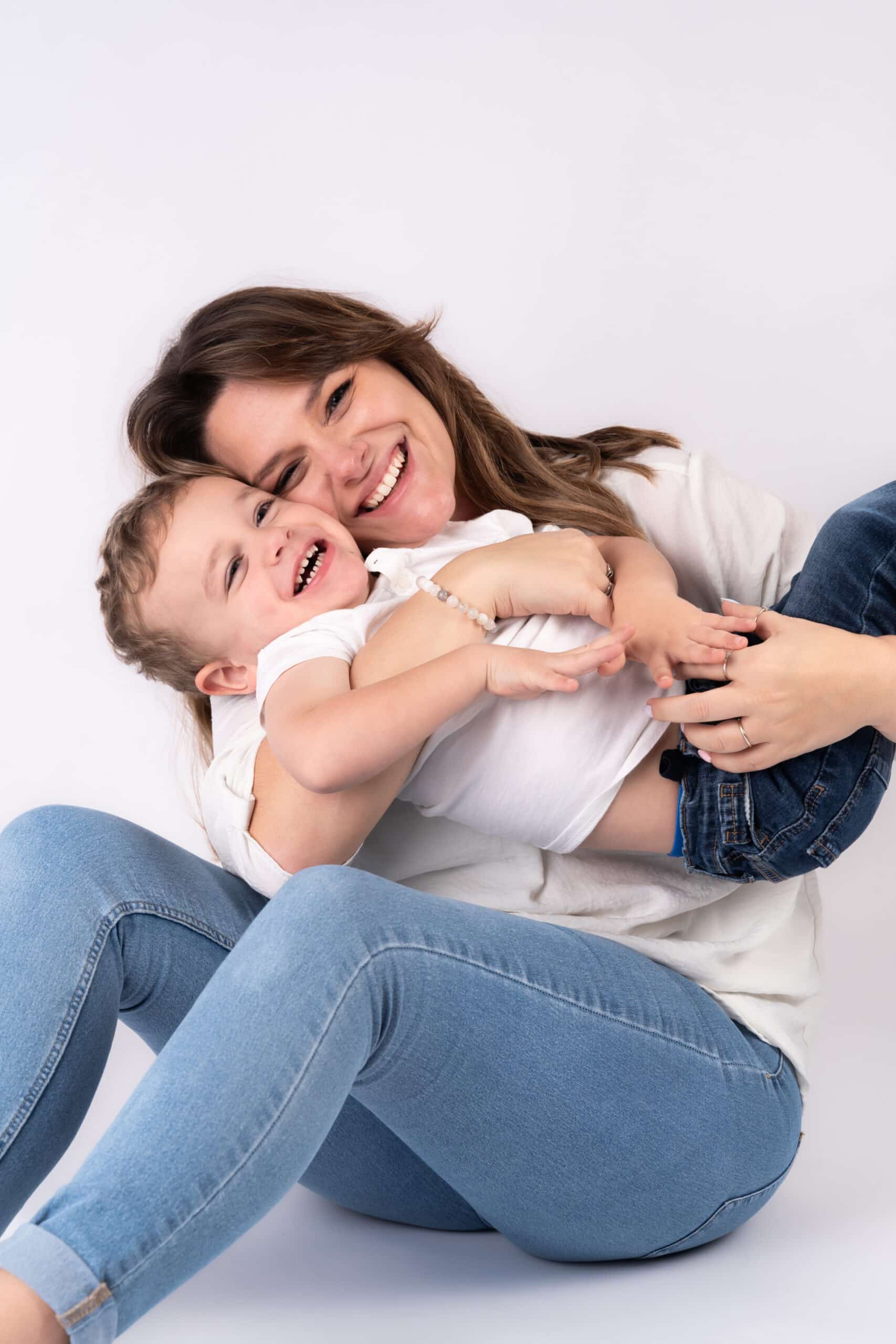 Une femme est assise par terre, les jambes croisées, tenant un enfant en bas âge qui rit sur ses genoux lors d'une délicieuse séance photo bébé. Tous deux portent des hauts blancs et des jeans bleus. La femme a les cheveux longs et sourit affectueusement tandis que le tout-petit rit, semblant joyeux et enjoué sur le fond blanc uni | Cette séance photo bébé est réalisée par Tribe Photography | Gaelle Massart Photographe