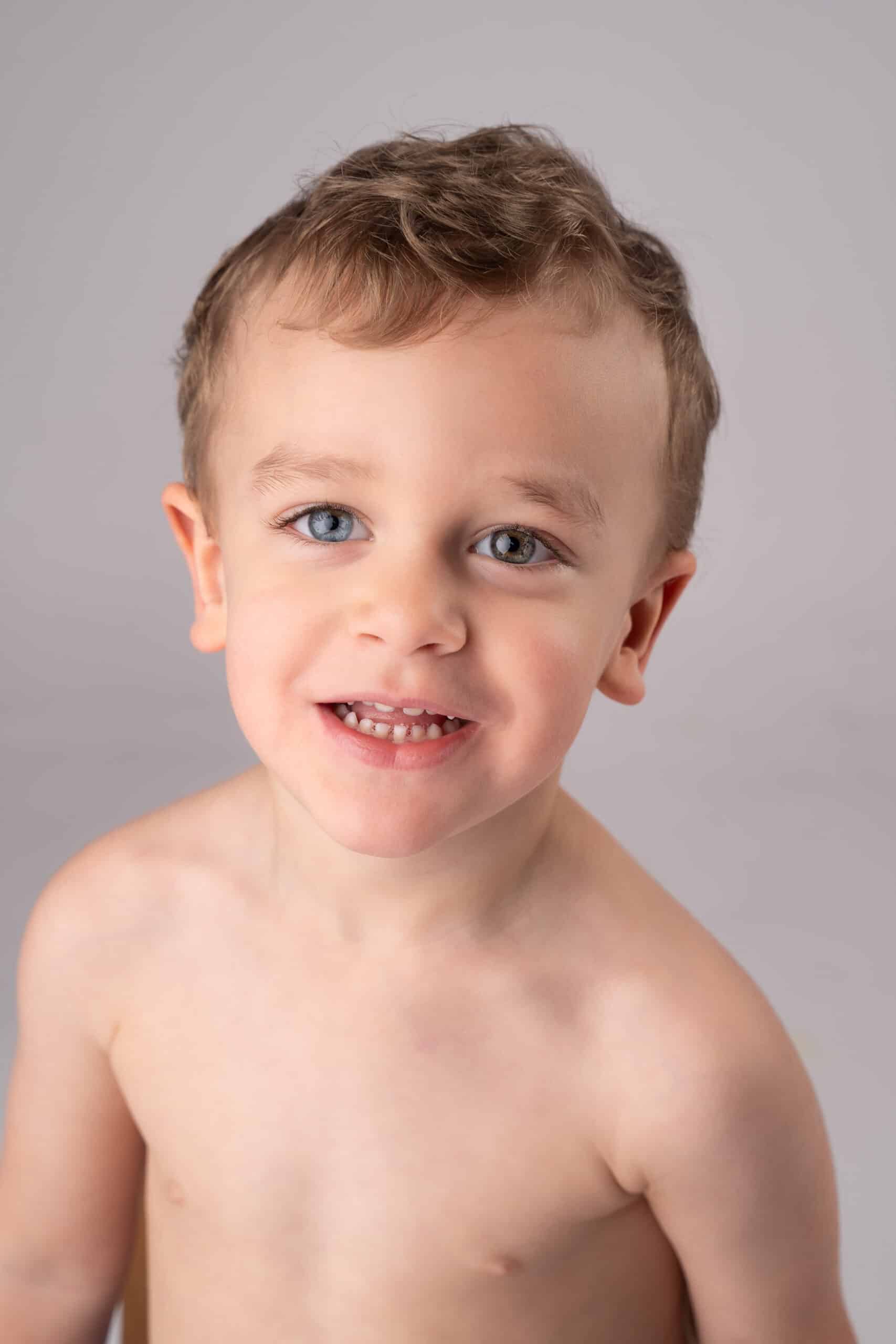 Un tout-petit aux cheveux courts châtain clair et aux yeux bleus sourit à l'appareil photo lors d'une séance photo bébé. L’enfant torse nu se tient sur un fond uni gris clair, capturant l’essence pure de l’enfance dans cette séance photo bébé | Ce shooting photo bébé est réalisé par Tribe Photography | Gaelle Massart Photographe