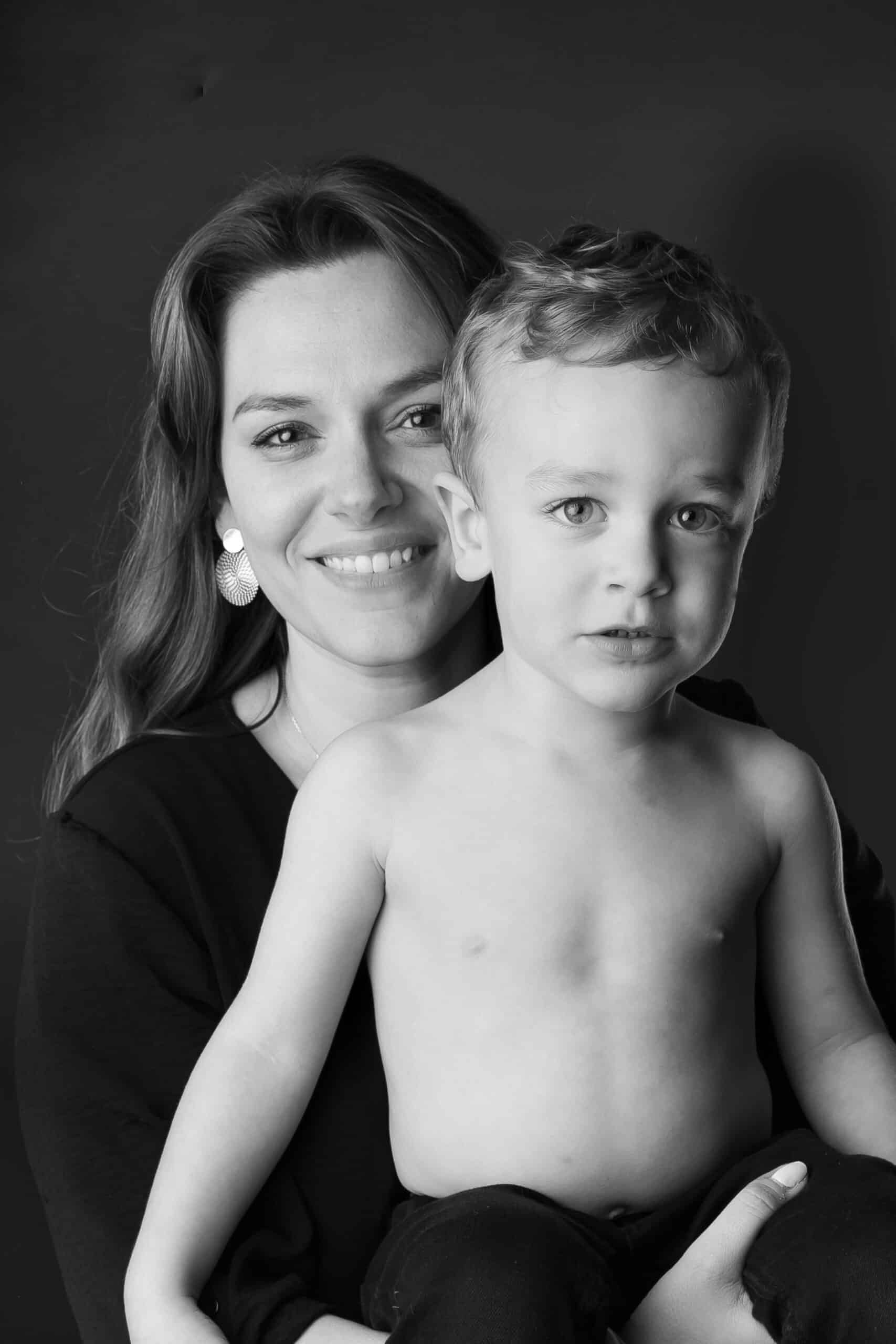 Une femme aux cheveux longs et aux grandes boucles d'oreilles, vêtue d'un haut sombre, sourit en tenant sur ses genoux un jeune enfant torse nu aux cheveux courts. Tous deux regardent la caméra, avec un fond sombre derrière eux. La photo est en noir et blanc, capturant le moment intime de leur séance photo bébé | Cette séance photo bébé est réalisée par Tribe Photography | Gaelle Massart Photographe