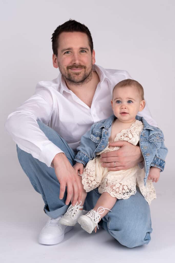 Un homme s'accroupit à  côté d'un bébé qu'il tient sur ses genoux lors d'une séance photo de famille. L'homme porte une chemise blanche, un jean bleu clair et des baskets blanches. Le bébé est habillé d'une robe en dentelle blanche et d'une veste en jean bleu clair, avec des petites chaussures blanches. Ils sourient tous les deux à  la caméra | Ce shooting photo famille est réalisé par Tribe Photography | Gaëlle Massart Photographe