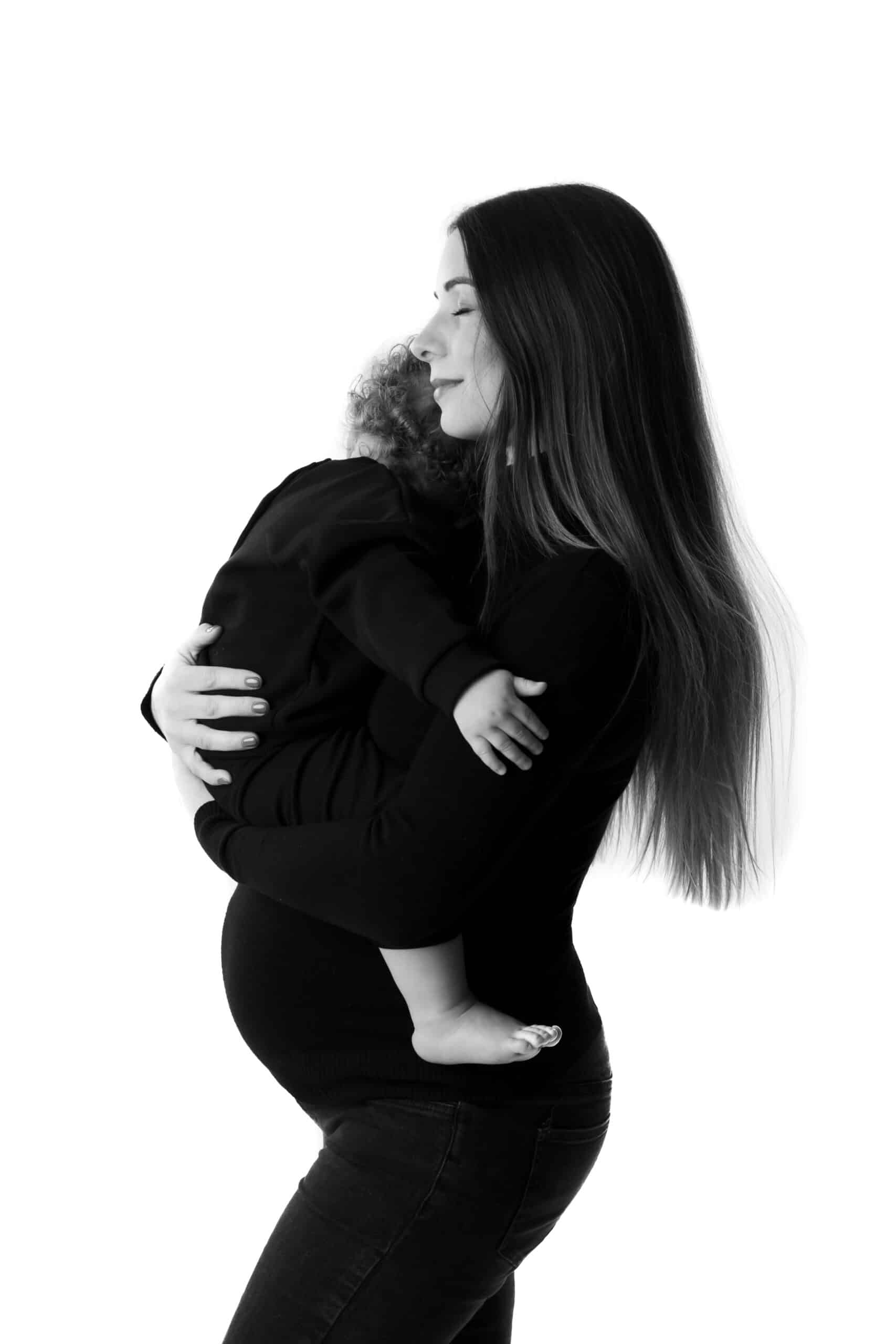 Une image en noir et blanc capture un moment poignant lors d’une séance photo bébé, mettant en vedette une femme aux cheveux longs tenant un petit enfant dans ses bras. La femme enceinte et l’enfant sont tous deux vêtus de vêtements sombres, sur un fond blanc uni | Cette séance photo bébé est réalisée par Tribe Photography | Gaelle Massart Photographe