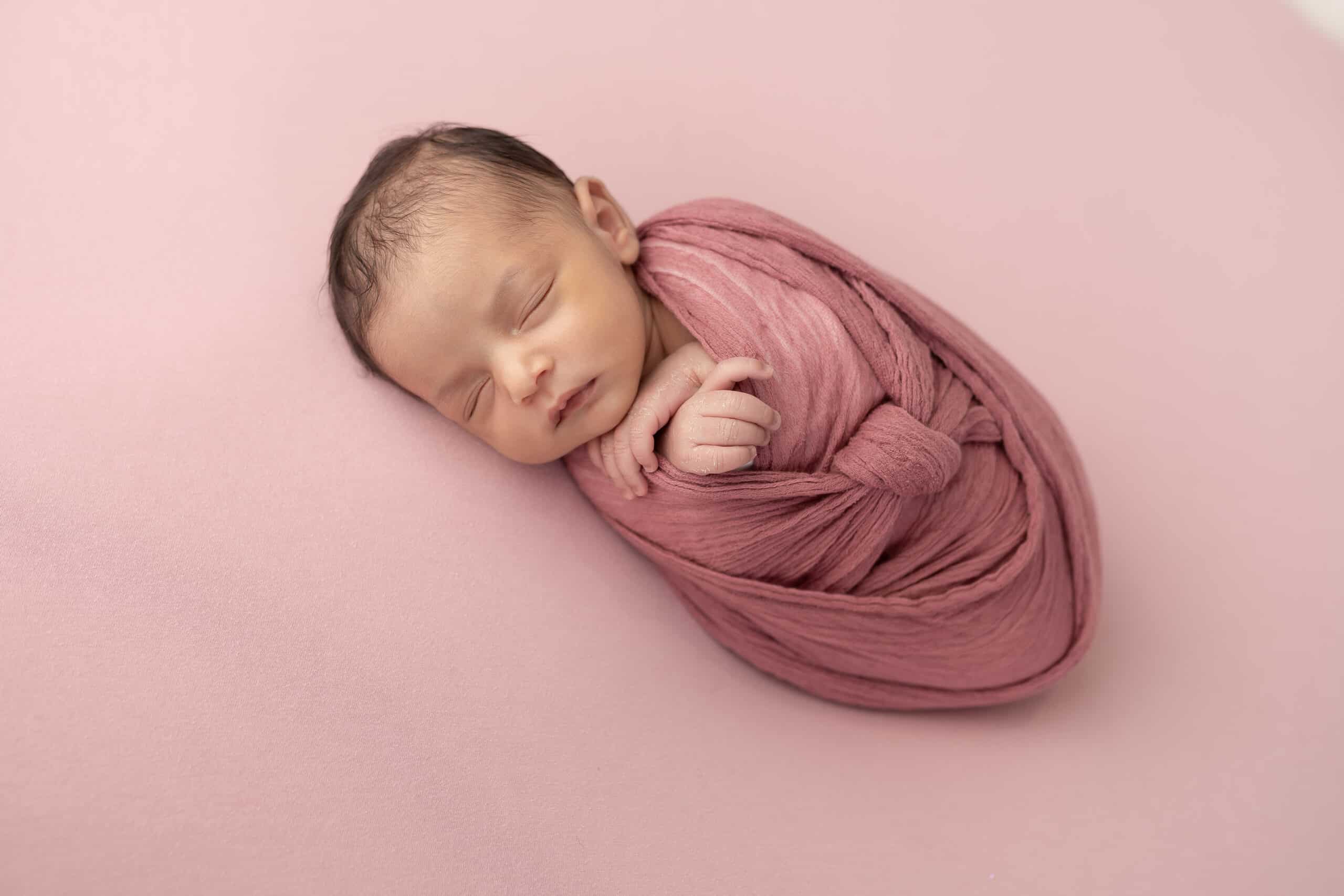 Un nouveau-né dort paisiblement emmailloté dans une couverture rose douce sur un fond rose assorti. La tête du bébé est légèrement tournée sur le côté, avec une petite main dépassant près de son visage. La scène respire la chaleur et la tranquillité, capturant l’essence d’une séance photo de naissance sereine | Ce shooting photo naissance est réalisé par Tribe Photography | Gaelle Massart Photographe