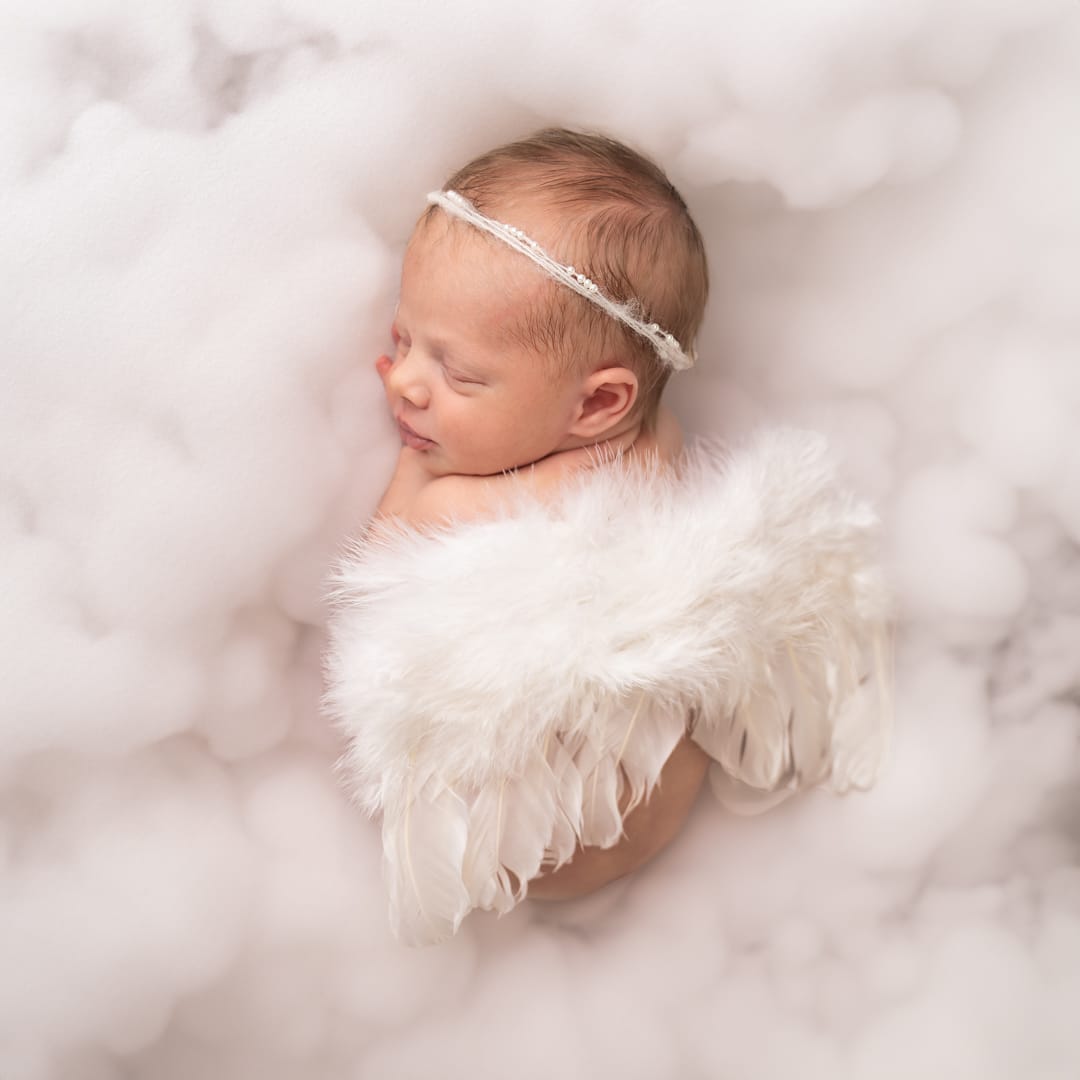 Un nouveau-né dort paisiblement sur une surface douce semblable à un nuage. Le bébé porte un bandeau délicat et des ailes d'ange à plumes blanches, ajoutant à l'atmosphère éthérée et sereine de la séance photo bébé fille | Ce shooting photo naissance fille est réalisé par Tribe Photography | Gaelle Massart Photographe