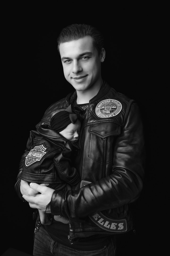 Image en noir et blanc d’un homme portant un gilet de club de moto en cuir, tenant dans ses bras un bébé endormi également vêtu d’une petite veste en cuir. L'homme sourit et regarde la caméra, debout sur un fond noir. Ce moment sincère capture l’esprit de leur séance photo de naissance spéciale | Ce shooting photo naissance est réalisé par Tribe Photography | Gaelle Massart Photographe