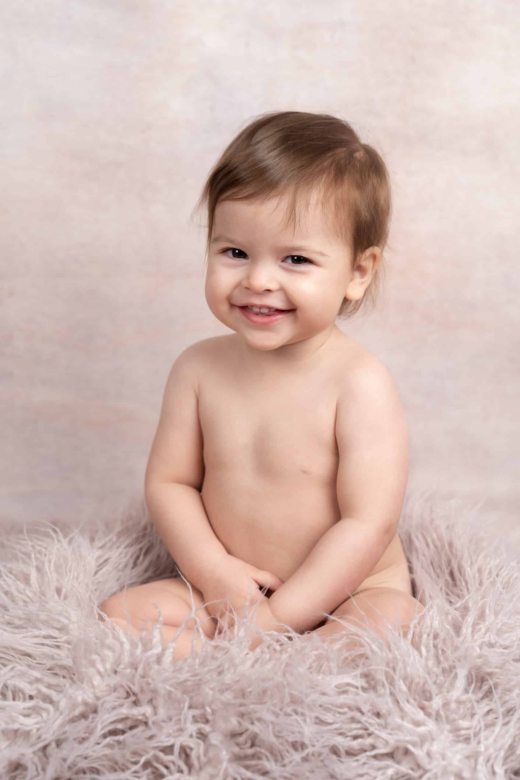 Un tout-petit souriant aux cheveux châtain clair est assis nu sur une surface douce et moelleuse sur un fond neutre. La prise de vue photo bébé capture le contentement et le bonheur de l'enfant, avec les mains doucement posées sur ses genoux | Cette photo de bébé est réalisée par Tribe Photography | Gaëlle Massart