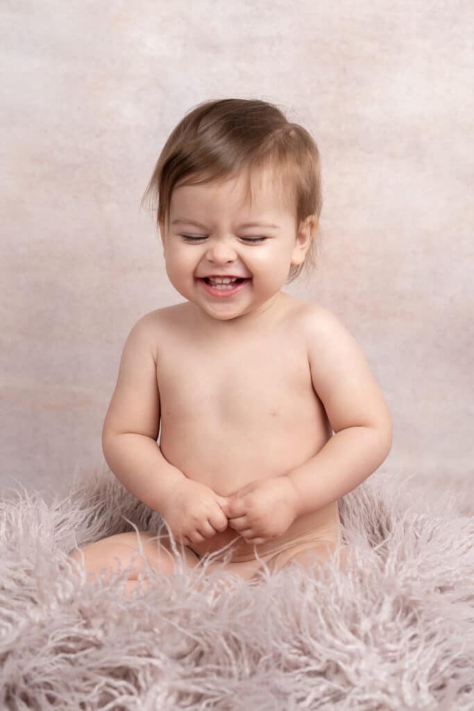 Un enfant en bas âge heureux, torse nu, aux cheveux bruns, est assis sur une couverture grise moelleuse sur un fond rose pâle et gris. L'enfant sourit, les yeux fermés et les mains jointes devant, parfait pour une charmante séance photo bébé garà§on | Cette photo de bébé est réalisée par Tribe Photography | Gaëlle Massart