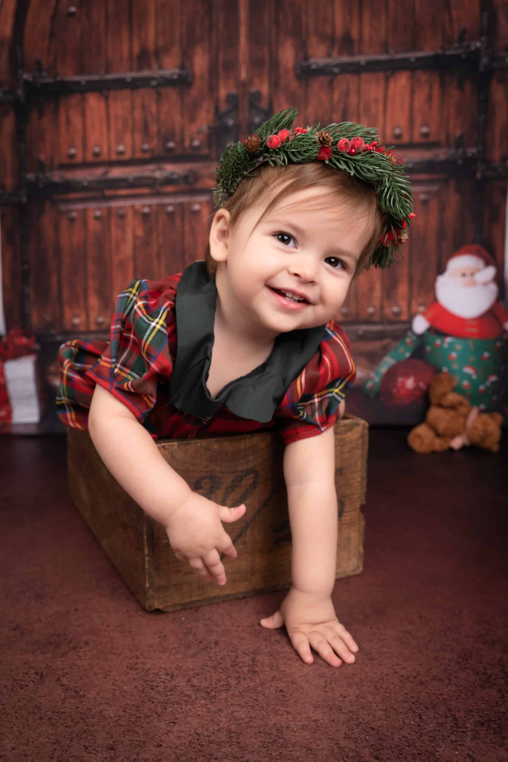 Un enfant souriant portant une robe à  carreaux et une couronne festive sur la tête est penché hors d'une boîte en bois. L'arrière-plan présente une porte en bois rustique, un décor de vacances avec une figurine du Père Noël et un ours en peluche. Cette photo bébé Noël capture magnifiquement l'esprit joyeux des fêtes | Ce shooting photo de Noël pour bébé est réalisée par Tribe Photography | Gaëlle Massart