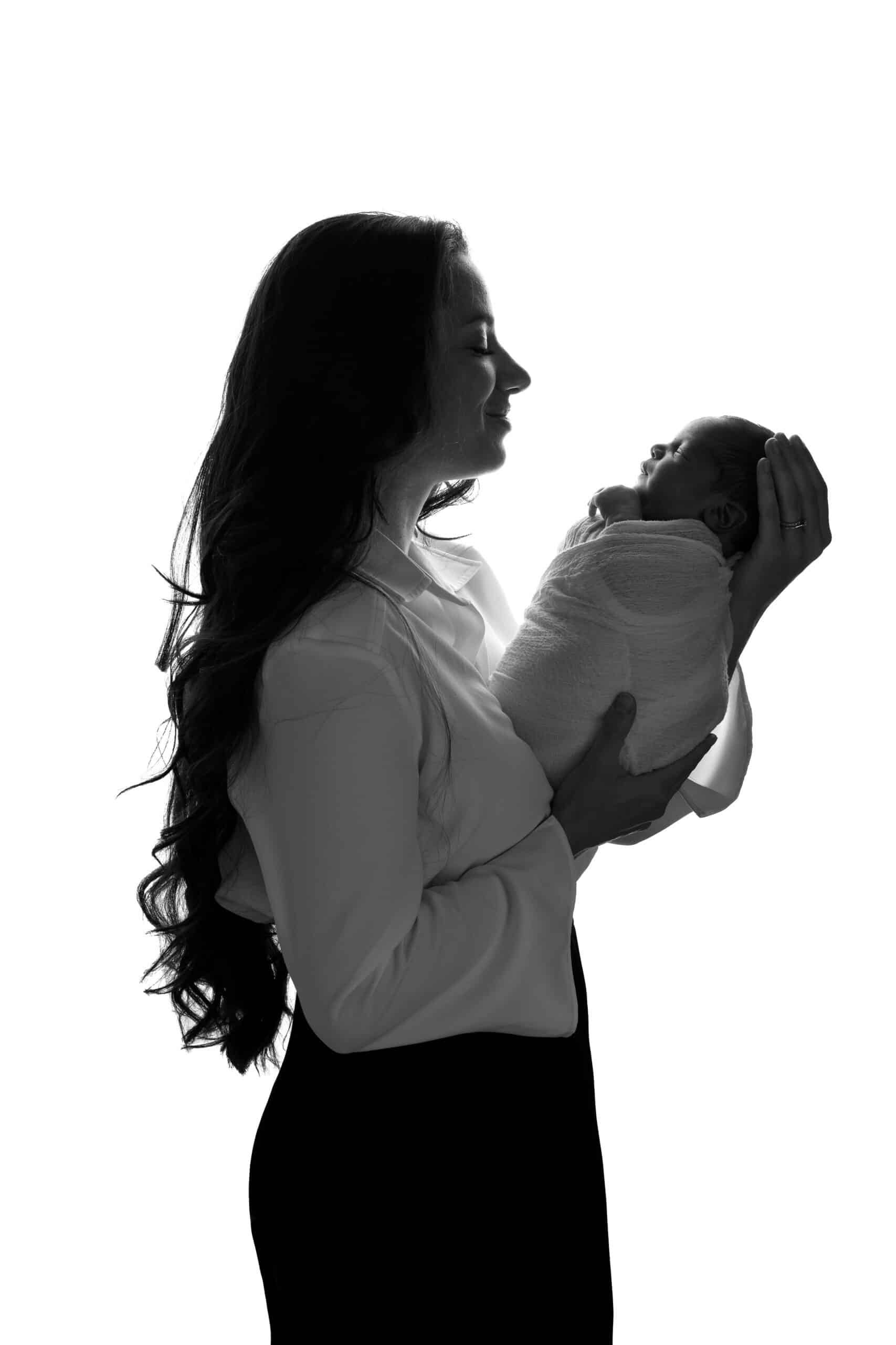 La silhouette d'une femme aux cheveux longs tenant un bébé emmailloté. Les deux se font face et le visage de la femme est légèrement tourné vers le bébé, créant un moment tendre et intime sur fond blanc, capturant l'essence d'une séance photo naissance | Ce shooting photo naissance est réalisé par Tribe Photography | Gaelle Massart Photographe