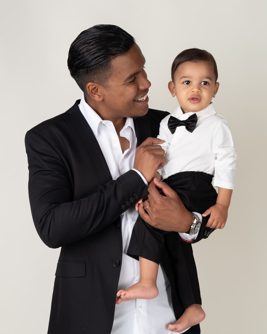 Un homme vêtu d'un costume noir et d'une chemise blanche tient dans ses bras un jeune enfant, vêtu d'un pantalon noir, d'une chemise blanche et d'un nœud papillon noir. Ils sourient tous les deux et regardent de côté lors d’une séance photo, sur un fond uni de couleur claire | Ce shooting photo bébé parents est réalisé par Tribe Photography | Gaelle Massart Photographe