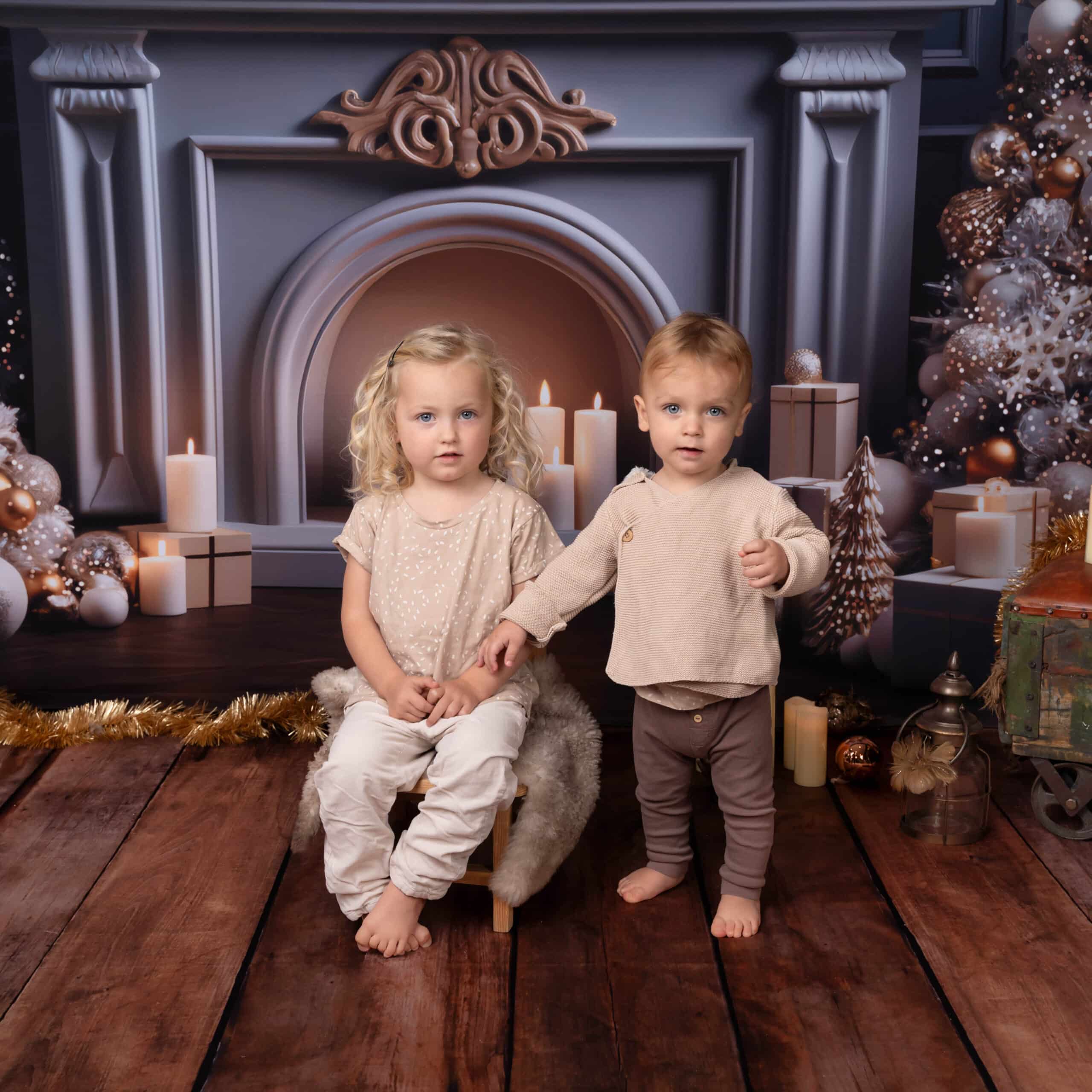 Deux jeunes enfants, une fille blonde assise et un garçon blond debout, tous deux vêtus de tenues beiges, posent pour une séance photo bébé Noël devant un décor festif mettant en vedette une cheminée décorée de bougies, de coffrets cadeaux et d'ornements. Le parquet est orné d'un tapis en fourrure à poils longs et de décorations de Noël | Ce shooting photo bébé Noël est réalisée par Tribe Photography | Gaelle Massart Photographe