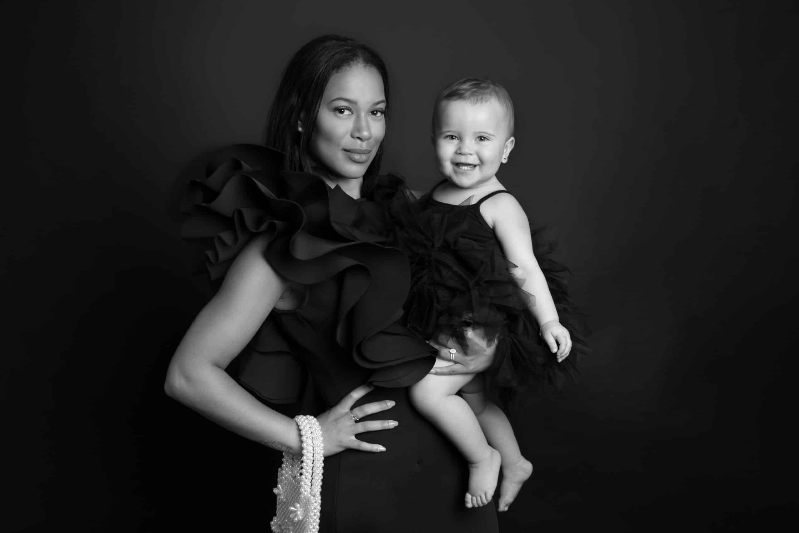 Photo en noir et blanc d'une femme et d'un bébé. La femme, vêtue d'une robe à  larges volants sur les manches, tient un rang de perles. Le bébé, vêtu d'une tenue à  volants, sourit tout en étant tenu par elle sur un fond sombre - un moment photo de bébé parfait, capturé de manière intemporelle | Cette séance photo bébé est réalisée par Tribe Photography | Gaëlle Massart Photographe