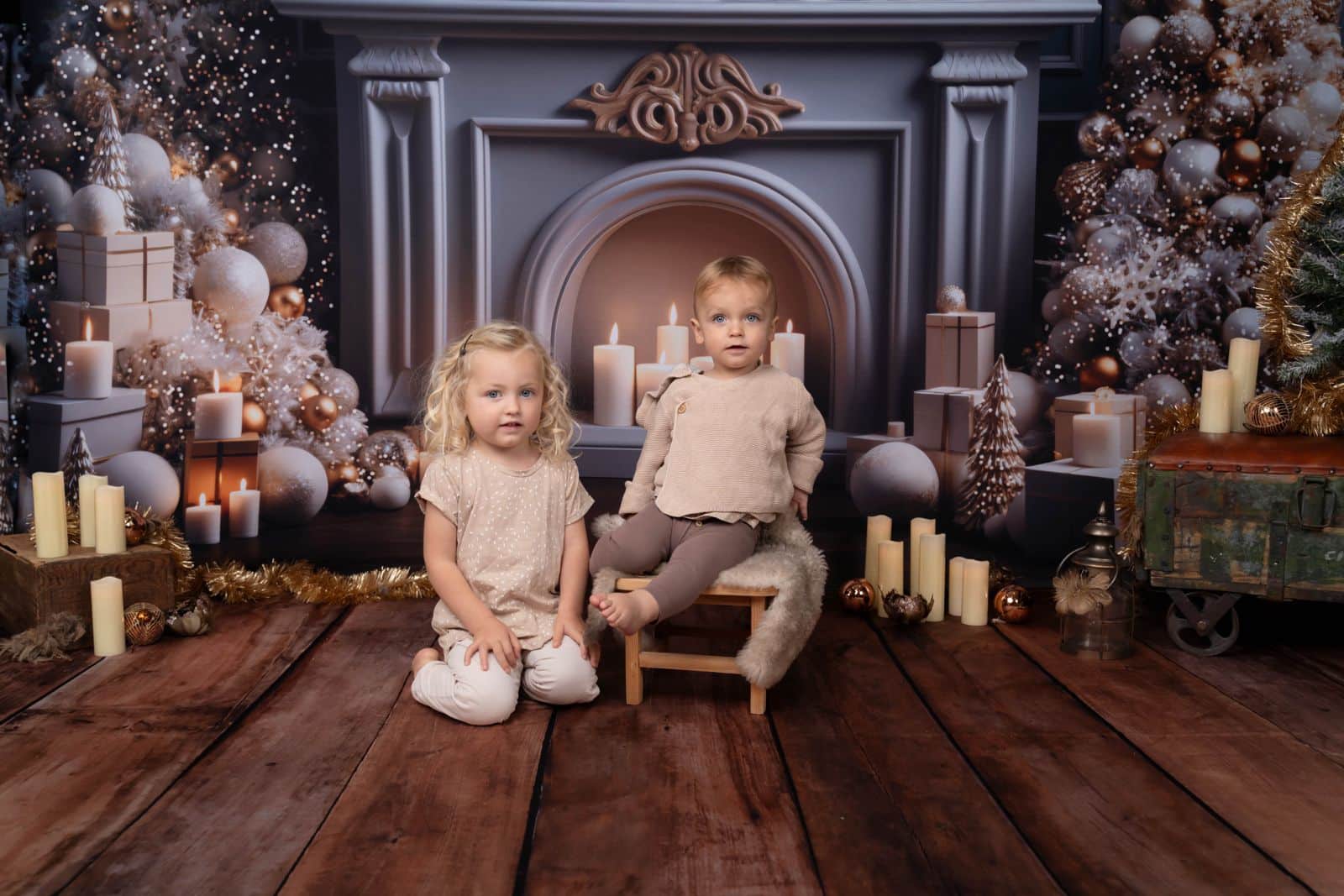 Deux jeunes enfants, une fille blonde assise et un garçon assis sur une chaise, tous deux vêtus de tenues beiges, posent pour une séance photo bébé Noël devant un décor festif mettant en vedette une cheminée décorée de bougies, de coffrets cadeaux et d'ornements. Le parquet est orné d'un tapis en fourrure à poils longs et de décorations de Noël | Ce shooting photo bébé Noël est réalisée par Tribe Photography | Gaelle Massart Photographe